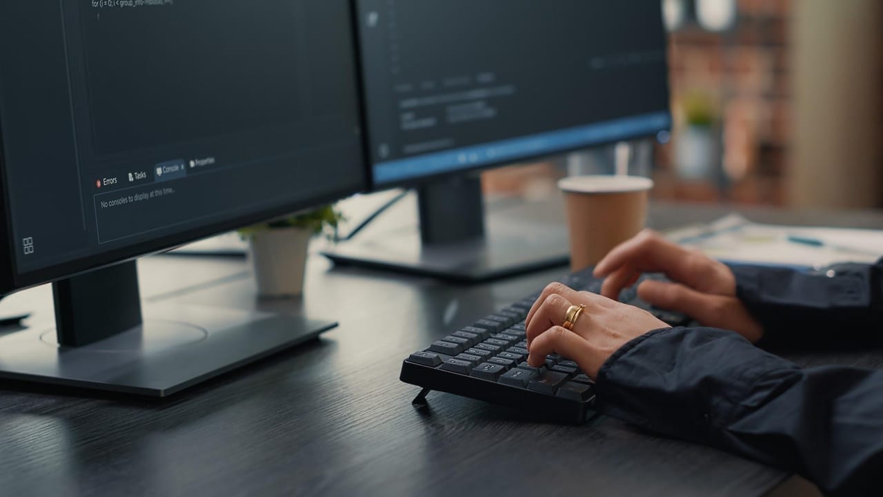 Hands at a keyboard, source code and a build console open on the monitors above.